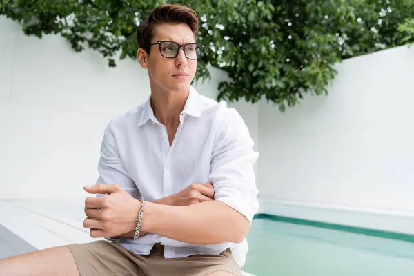 man with silver bracelet and eyeglasses sitting near pool and looking away