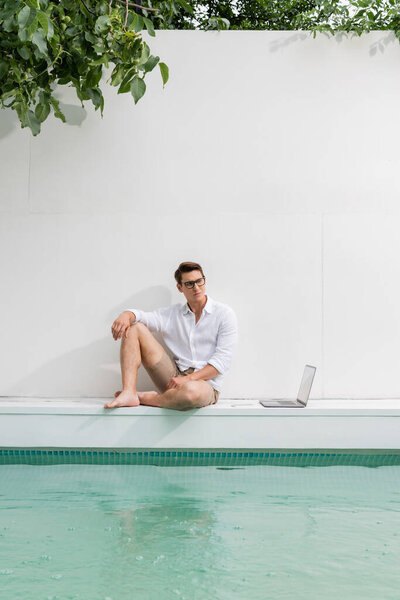 полная длина босиком человек в очках, сидя у бассейна рядом с белой стеной и ноутбуком