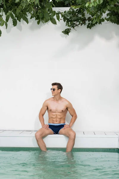 smiling man in sunglasses and swimming trunks sitting near pool and looking away