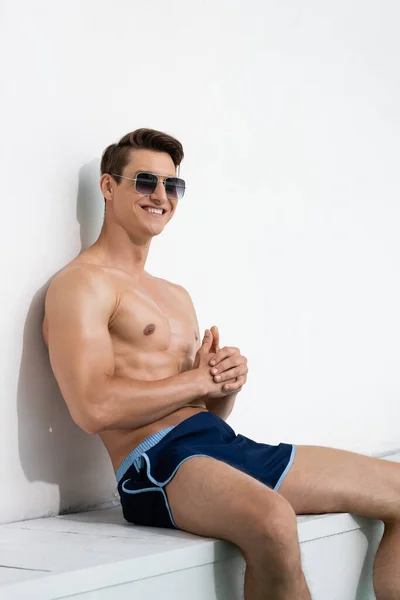 Cheerful Athletic Man Sunglasses Swimwear Sitting White Wall — Stockfoto