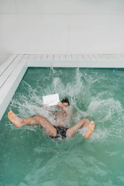high angle view of man with laptop falling in pool with water splashes