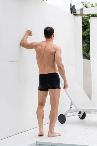 back view of wet muscular man in swimming trunks near white wall and deck chair