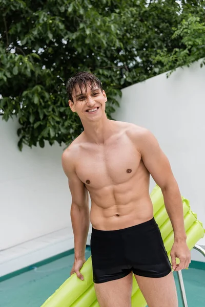 happy muscular man with wet hair holding inflatable mattress at poolside