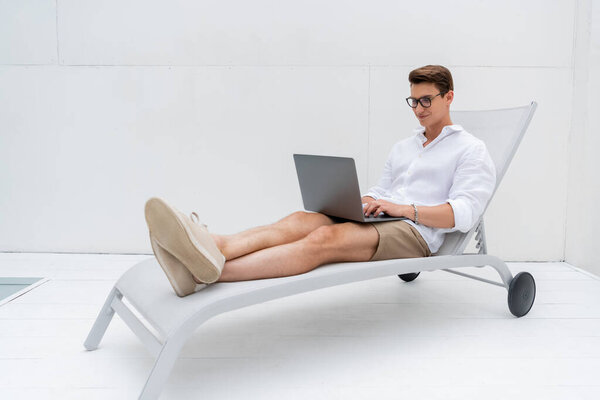 full length of smiling freelancer in summer clothes sitting in deck chair and typing on laptop