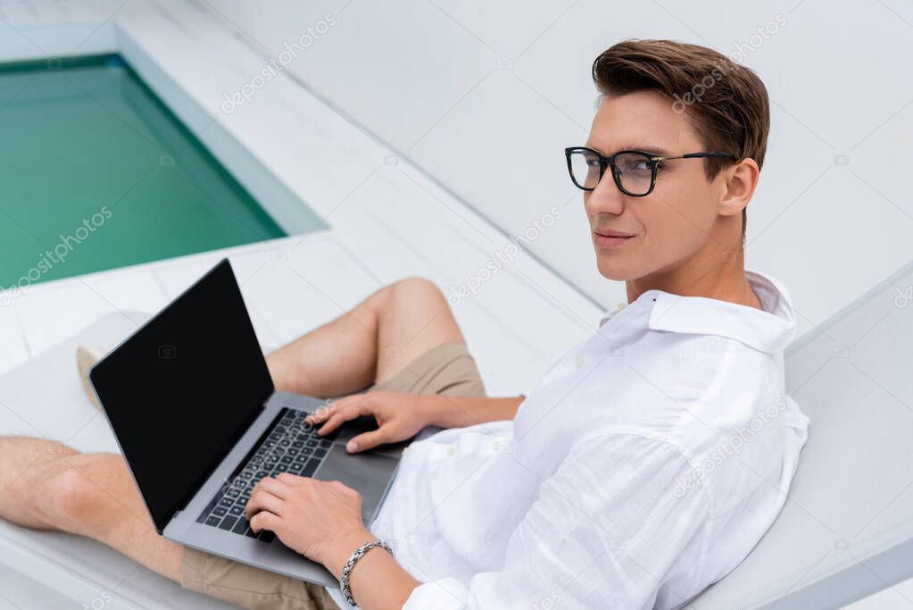 man in eyeglasses looking at camera near laptop with blank screen in deck chair