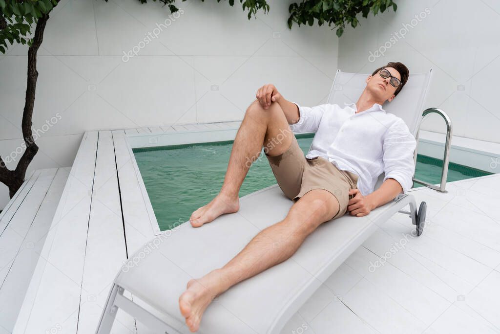 barefoot man in shorts and eyeglasses lying on deck chair near pool