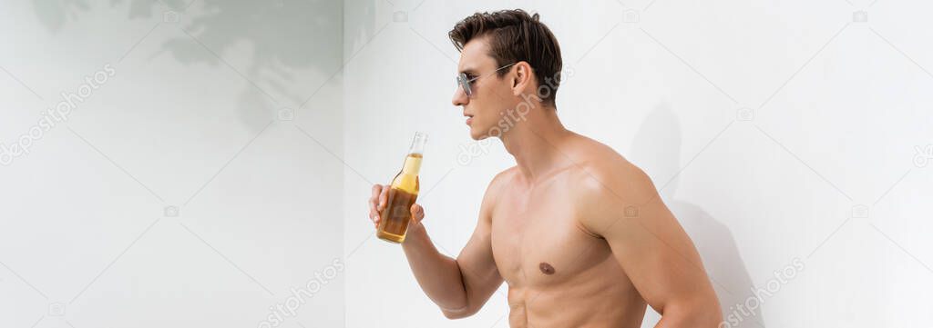 shirtless and muscular man in sunglasses drinking beer near white wall, banner