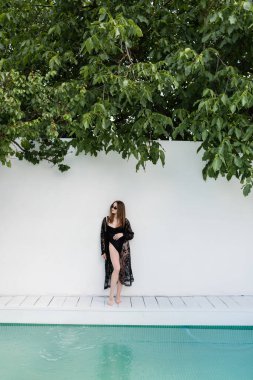 Mayo giymiş şık bir kadın duvarın yanında duruyor ve havuz başında. 