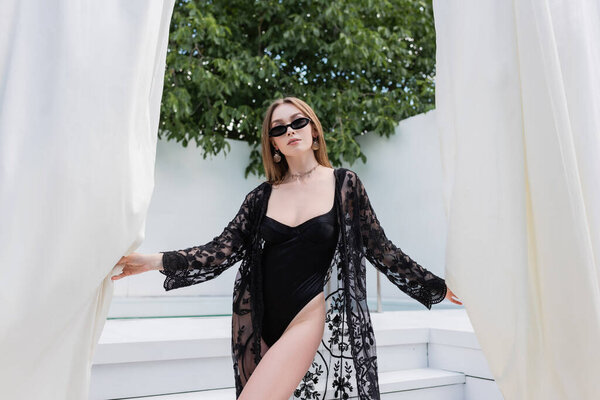Trendy woman in swimsuit and guipure robe holding curtains at resort 