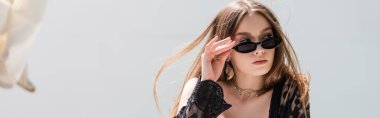 Stylish woman in sunglasses and guipure robe holding sunglasses at resort, banner 