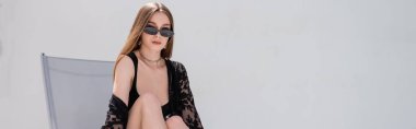 Guipure bornoz ve güneş gözlüğü takmış şık bir kadın tatil köyünde iskemlede oturuyor. 