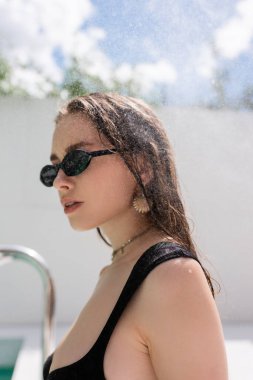 Trendy young woman in sunglasses standing under water drops at resort 