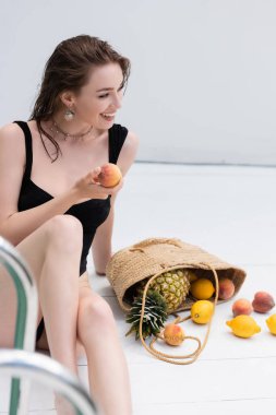 Mayo giymiş gülümseyen kadın tatil köyünde saman çantasındaki meyvelerin yanında taze şeftali tutuyor. 