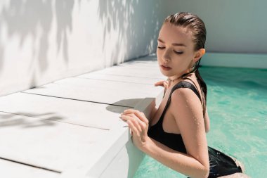 pretty young woman with wet hair and stylish black swimsuit in pool 