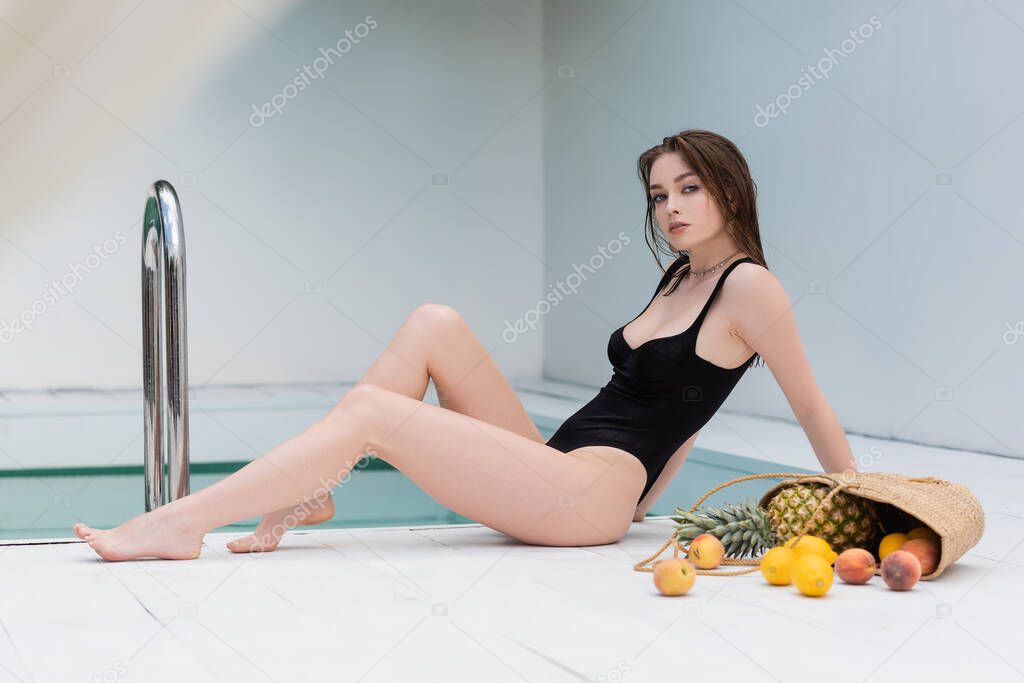 Pretty young woman in swimsuit sitting near tropical fruits and pool at resort 