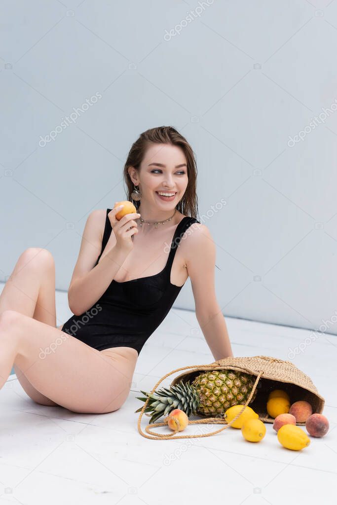 Cheerful young woman in swimsuit holding peach near fruits in straw handbag at resort 