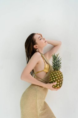 Duvarın yanında olgun ananas tutan altın mayo ve elbise giymiş gülümseyen bir kadın. 