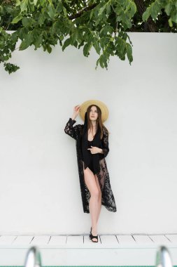 Beyaz duvarın yanında siyah örgü bornozla dikilmiş hasır şapka ayarlayan zarif genç bir kadın. 