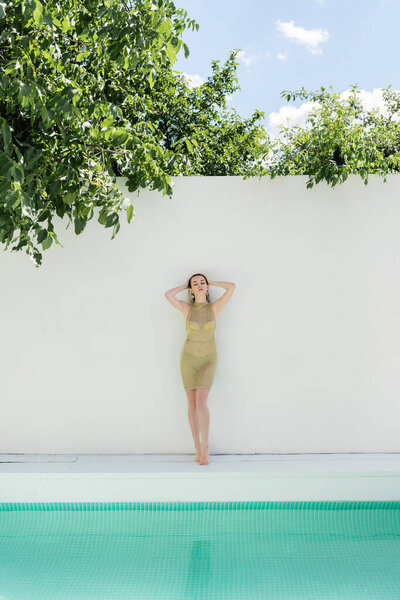 full length of barefoot young woman in golden summer dress standing near while wall and swimming pool 