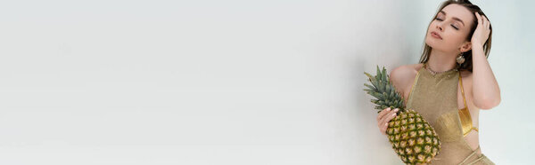 young woman in golden summer dress holding fresh pineapple near white wall, banner