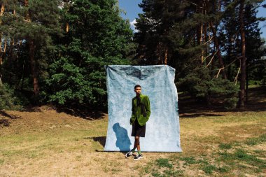 full length of african american man in black shorts and green blazer near blue drape in park