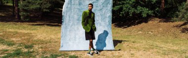 full length of african american man in green blazer and black shorts standing near blue cloth in forest, banner