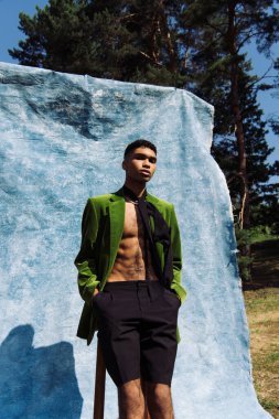 african american man in green blazer and neck scarf standing with hands in pockets near blue cloth in park
