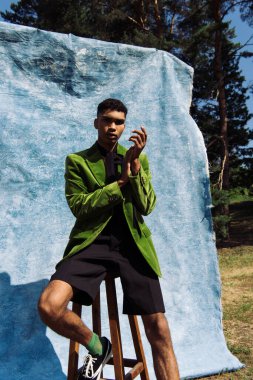 trendy african american man in green blazer and shorts looking at camera while sitting near blue cloth outdoors