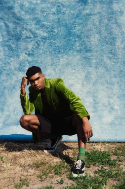full length of african american man in green blazer and sneakers posing on haunches near blue drapery 