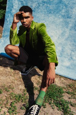 african american man in green blazer and sneakers looking at camera near blue cloth outdoors