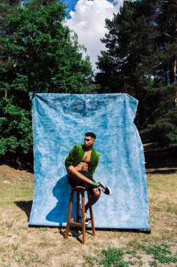 full length of african american man in black shorts and green blazer posing on stool in forest near blue cloth