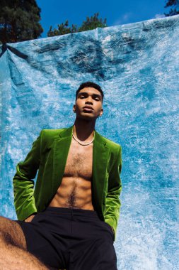 low angle view of african american man in green blazer posing with hands in pockets near blue cloth