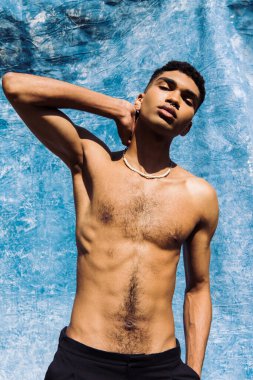 Boncuklu genç Afro-Amerikan adam mavi perdenin yanında eli boynunun arkasında poz veriyor.