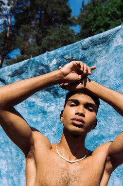 low angle view of shirtless african american in beads posing near blue cloth outdoors