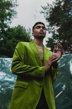 low angle view of african american man in green blazer and beads looking at camera near mirror in park
