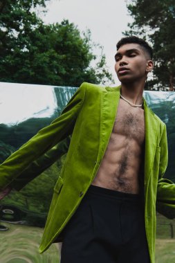young african american man in green blazer and beads posing near mirror in park