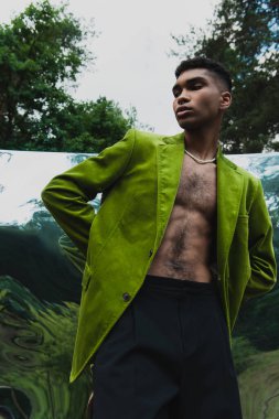 trendy african american man in green blazer standing with hands behind back near mirror surface in forest