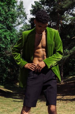 young african american man in black shorts and green blazer standing with hand on hip in forest
