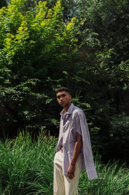 young african american man in blue and striped shirt looking at camera in summer park