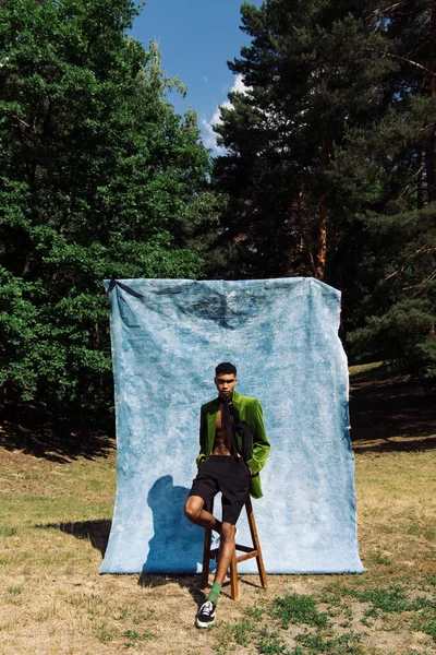 trendy african american man in blazer and shorts posing with hands in pockets near stool and blue drape