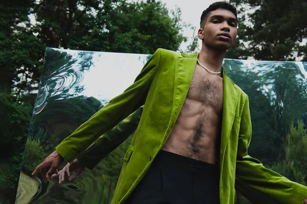 low angle view of african american man in fashionable blazer posing near mirror in forest