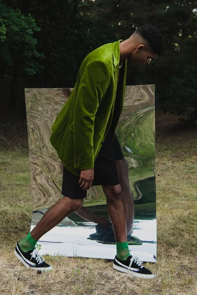 side view of african american man in green velvet blazer and shorts walking near flexible mirror in park