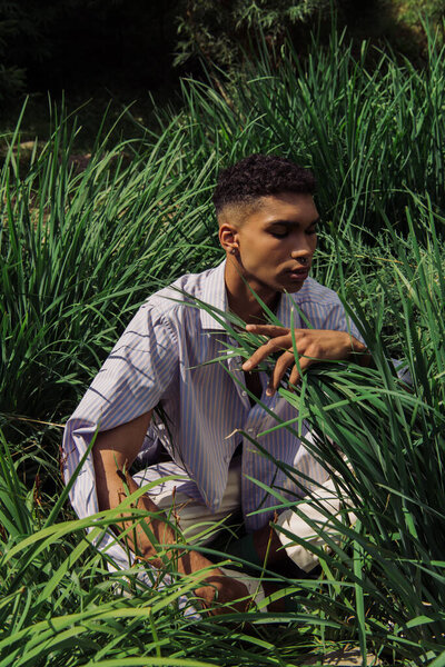 young african american man in blue and striped shirt sitting on grass in park