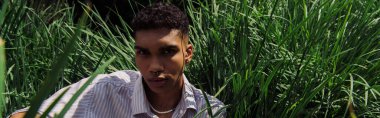 young african american man with piercing looking at camera near green grass, banner