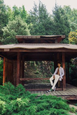full length of trendy african american man in wooden gazebo in green park