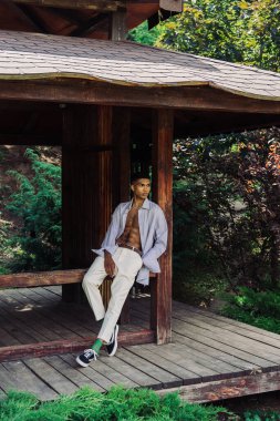 african american man in white trousers and blue shirt posing on wooden terrace in park