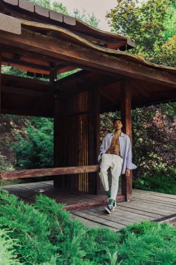 young african american man in stylish clothes posing in wooden gazebo in green park