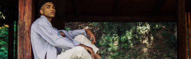 stylish african american man in striped shirt sitting in wooden gazebo and looking at camera, banner