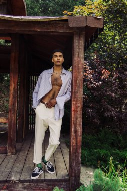 full length of african american man in stylish clothes looking at camera in wooden gazebo