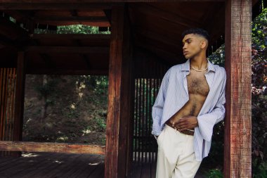 trendy african american man in blue shirt posing in park in wooden gazebo